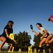 Two women and a man playing Spikeball with paddles on a grassy field under a clear blue sky. The players are actively hitting the yellow ball, and a Spikeball net is set up on the ground.