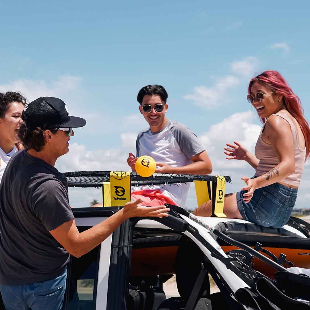 Four friends gather around a car with a Spikeball net set up on the roof. They are smiling and laughing under a clear blue sky, with one person seated on the roof holding a Spikeball.